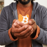 Primer plano de un hombre con vestimenta casual sosteniendo con ambas manos una pequeña alcancía naranja con el logotipo de Bitcoin, simbolizando la protección y la autocustodia de activos digitales en un entorno doméstico.
