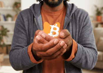 Primer plano de un hombre con vestimenta casual sosteniendo con ambas manos una pequeña alcancía naranja con el logotipo de Bitcoin, simbolizando la protección y la autocustodia de activos digitales en un entorno doméstico.
