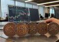 Cinco monedas de bronce con los logos de DODO, HEI, HFT, SYN y TLM alineadas sobre un escritorio frente a una pantalla con gráficos financieros de Binance.