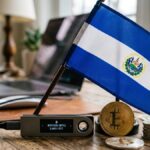 Una hardware wallet con bitcoin y la bandera de El Salvador sobre un escritorio.