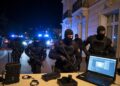 Operación policial en España varios agentes de la Policía Nacional con chalecos antibalas y pasamontañas irrumpiendo en una vivienda de lujo durante la noche. Luces azules de patrullas policiales iluminando la escena.