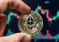 Mano sosteniendo una moneda física de Ethereum con gráficos bursátiles en el fondo.