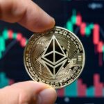 Mano sosteniendo una moneda física de Ethereum con gráficos bursátiles en el fondo.
