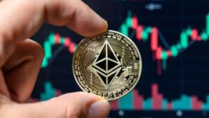 Mano sosteniendo una moneda física de Ethereum con gráficos bursátiles en el fondo.