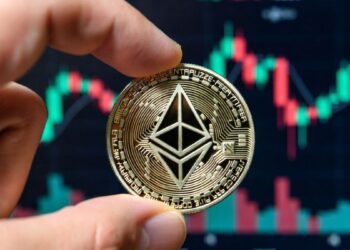 Mano sosteniendo una moneda física de Ethereum con gráficos bursátiles en el fondo.