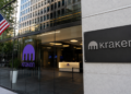 Entrada de oficinas corporativas de Kraken con fachada de vidrio, logotipo de la empresa y una bandera de Estados Unidos en el exterior