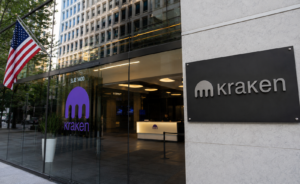 Entrada de oficinas corporativas de Kraken con fachada de vidrio, logotipo de la empresa y una bandera de Estados Unidos en el exterior