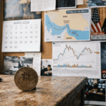 En primer plano: moneda de bitcoin sobre una mesa. En el fondo: imágenes de guerra en irán, un calendario de mayo de 2026 y una foto del presidente Trump hablando.
