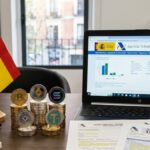 Criptomonedas con la bandera de España y una laptop que muestra el portal de la Agencia Tributaria, con anteojos, papeles y lapiceros, sobre un escritorio.