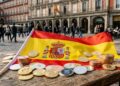Criptomonedas alrededor de la bandera de España sobre una mesa de madera en una plaza de Madrid.