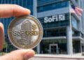 Mano sosteniendo una moneda física con la inscripción "SoFiUSD" en primer plano frente a un edificio moderno de SoFi Bank.