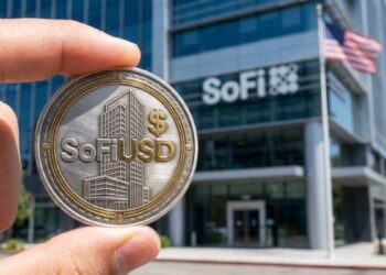 Mano sosteniendo una moneda física con la inscripción "SoFiUSD" en primer plano frente a un edificio moderno de SoFi Bank.
