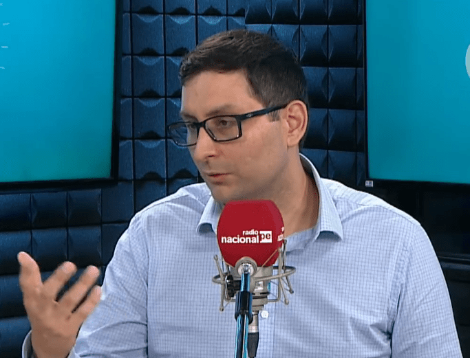 Fotografía en plano medio de Adolfo Morán durante una entrevista en una cabina de radio. Viste una camisa clara y lleva anteojos. Está hablando con un gesto reflexivo en su mano derecha frente a un micrófono profesional de mesa que lleva el logotipo rojo de "Radio Nacional".