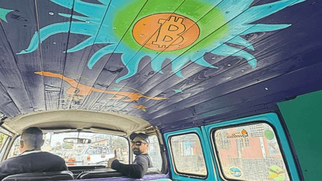 Interior de una furgoneta Volkswagen clásica tomada desde el asiento trasero. El techo del vehículo está revestido con tablones de madera pintados de morado, donde destaca un gran sol artístico en tonos turquesa y verde brillante con el logo de Bitcoin en color naranja en el centro.