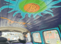 Interior de una furgoneta Volkswagen clásica tomada desde el asiento trasero. El techo del vehículo está revestido con tablones de madera pintados de morado, donde destaca un gran sol artístico en tonos turquesa y verde brillante con el logo de Bitcoin en color naranja en el centro.