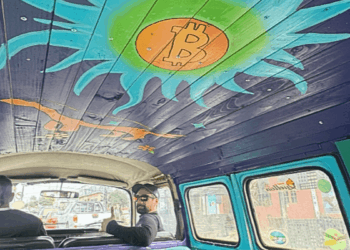 Interior de una furgoneta Volkswagen clásica tomada desde el asiento trasero. El techo del vehículo está revestido con tablones de madera pintados de morado, donde destaca un gran sol artístico en tonos turquesa y verde brillante con el logo de Bitcoin en color naranja en el centro.
