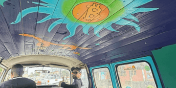 Interior de una furgoneta Volkswagen clásica tomada desde el asiento trasero. El techo del vehículo está revestido con tablones de madera pintados de morado, donde destaca un gran sol artístico en tonos turquesa y verde brillante con el logo de Bitcoin en color naranja en el centro.