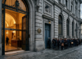 Una fachada institucional de piedra, sobria, de banco europeo clásico, con dos puertas contiguas: una ancha, monumental, abierta de par en par, con luz cálida saliendo de ella; al lado, una puerta angosta, baja, con una larga fila difusa esperando frente a ella. Sin texto. Paleta institucional fría —grises piedra, azul europeo apagado, dorado tenue en la luz de la puerta ancha—. Iluminación de media tarde, sombras largas. Composición horizontal 16:9, la puerta ancha desplazada a un tercio, espacio negativo arriba. Estética editorial, fotográfica, seria, sin caricatura.