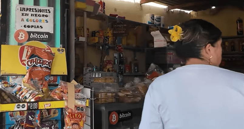 Interior de una pequeña bodega o comercio minorista local en Perú. En primer plano, de espaldas, se observa a una mujer con una flor amarilla en el cabello frente al mostrador. A la izquierda, hay estantes con productos como snacks (Chizitos, Lays). Destacan de manera visible dos stickers con el logotipo naranja de Bitcoin y la frase "Aceptamos aquí",