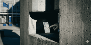 Fotografía conceptual de estilo editorial financiero. Una imponente y fría estructura de hormigón, que evoca la arquitectura institucional de Bruselas, proyecta una sombra densa y afilada sobre la penumbra. Atrapada en el centro de esa sombra, la pantalla de un teléfono móvil brilla tenuemente, mostrando la interfaz limpia de un gráfico financiero de velas en caída libre y el símbolo de la moneda estable Tether (USDT).