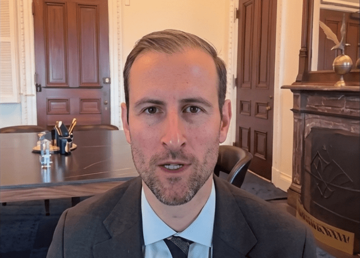 Primer plano de Patrick Witt durante una transmisión de video en vivo. Viste traje oscuro, camisa blanca y corbata, y aparece hablando frente a la cámara. Al fondo se observa una sala de reuniones con una mesa grande, una puerta de madera de estilo clásico.
