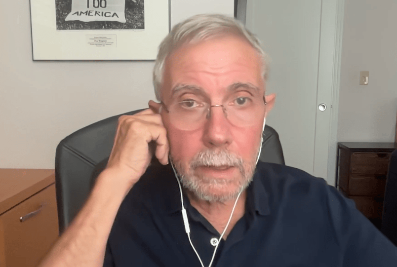 Retrato de medio cuerpo del economista estadounidense Paul Krugman, premio Nobel de Economía, hablando de manera analítica en una conferencia. Se le observa de frente con una expresión seria y gesticulando levemente, vestido con una camisa oscura sobre un fondo de oficina.