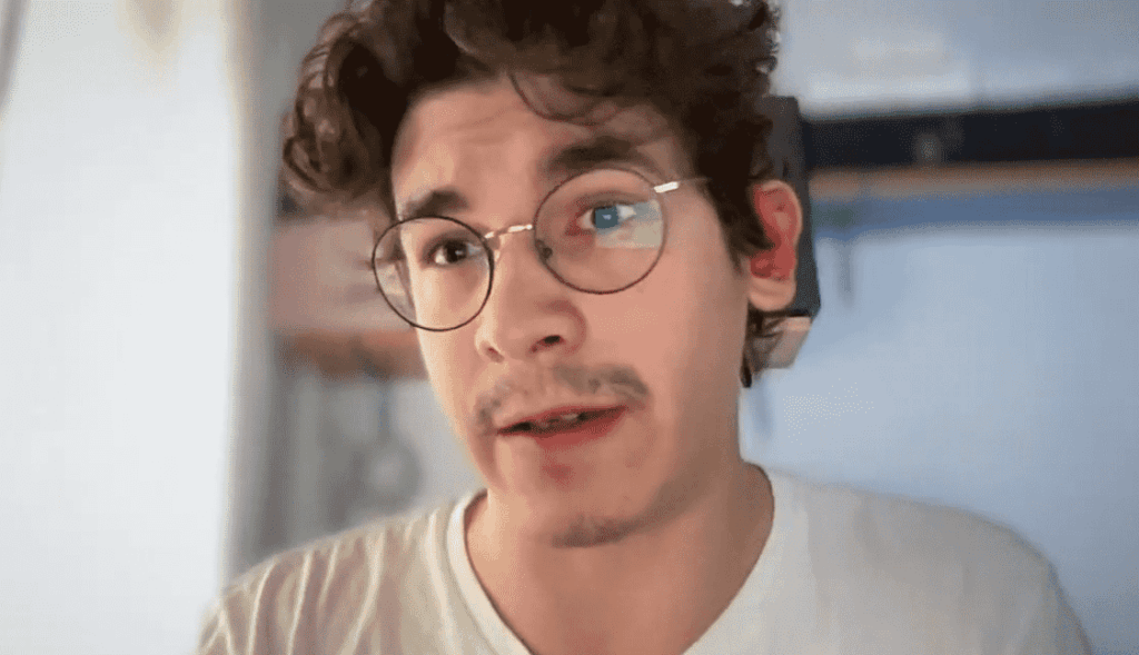Primer plano de Rodrigo Rivas durante una videollamada con CriptoNoticias. Se observa a un hombre joven de cabello castaño y rizado, con anteojos redondos de montura metálica delgada y un bigote ligero. Viste una camiseta blanca y aparece hablando de frente a la cámara, con un fondo interior sutilmente difuminado.