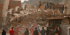 Plano medio general a plena luz del día que muestra el colapso total de un edificio residencial de varias plantas. La estructura ha quedado reducida a enormes losas de hormigón armado apiladas entre escombros y polvo. Decenas de rescatistas y miembros de cuerpos de emergencia en Venezuela.