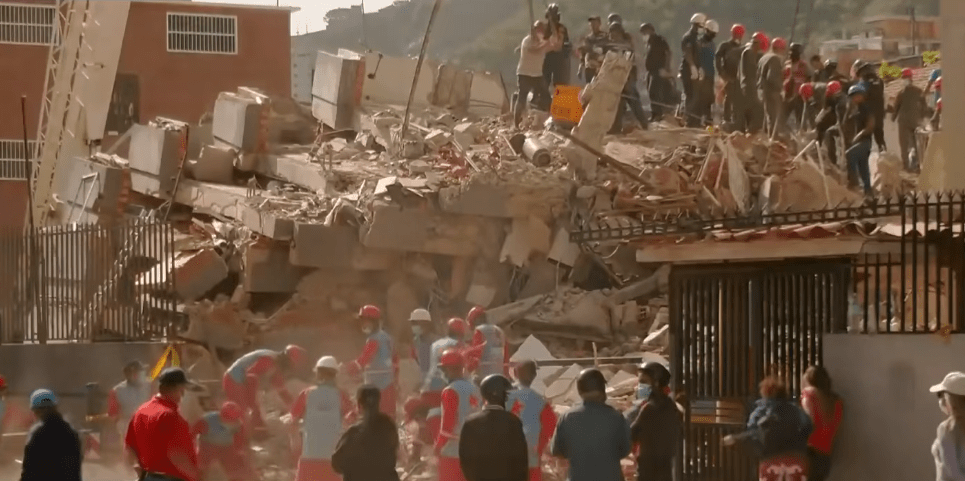 Plano medio general a plena luz del día que muestra el colapso total de un edificio residencial de varias plantas. La estructura ha quedado reducida a enormes losas de hormigón armado apiladas entre escombros y polvo. Decenas de rescatistas y miembros de cuerpos de emergencia en Venezuela.