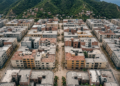 Fotografía aérea editorial, formato horizontal. Vista desde altura de un barrio urbano latinoamericano densamente edificado tras un terremoto: edificios de concreto con fachadas agrietadas o parcialmente derrumbadas, calles cubiertas de escombros y polvo, sin vegetación. Composición geométrica con las líneas de las calles y los bloques de edificios como estructura visual. Paleta desaturada de grises, beige y ocre terroso. Luz de día nublado, sin sombras duras. Atmósfera documental y fría, sin dramatismo exagerado. Sin personas visibles, sin texto, sin números, sin logotipos ni marcas.