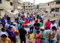 Imagen horizontal realista estilo fotográfico. Escena neutral de ayuda humanitaria tras terremoto en Venezuela: edificios colapsados, escombros, voluntarios comunitarios distribuyendo comida, agua y mantas a familias afectadas en una calle urbana.