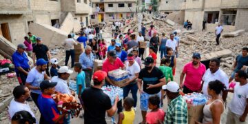 Imagen horizontal realista estilo fotográfico. Escena neutral de ayuda humanitaria tras terremoto en Venezuela: edificios colapsados, escombros, voluntarios comunitarios distribuyendo comida, agua y mantas a familias afectadas en una calle urbana.