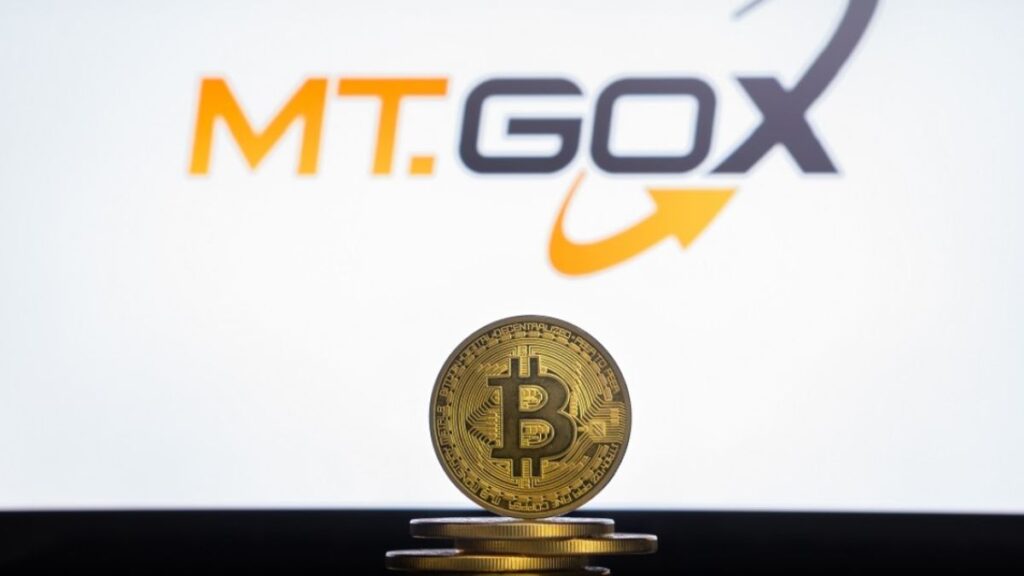 Moneda física de bitcoin dorada sobre un escritorio con el logo de Mt.Gox de fondo.