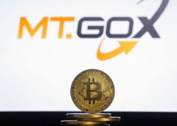 Moneda física de bitcoin dorada sobre un escritorio con el logo de Mt.Gox de fondo.