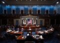 Imagen realista horizontal estilo fotografía periodística. En el interior del Senado de Estados Unidos, con la cámara del Senado visible, senadores sentados en sus escritorios de madera oscura mientras votan. En primer plano, una mano presionando un botón de votación electrónica.