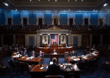 Imagen realista horizontal estilo fotografía periodística. En el interior del Senado de Estados Unidos, con la cámara del Senado visible, senadores sentados en sus escritorios de madera oscura mientras votan. En primer plano, una mano presionando un botón de votación electrónica.