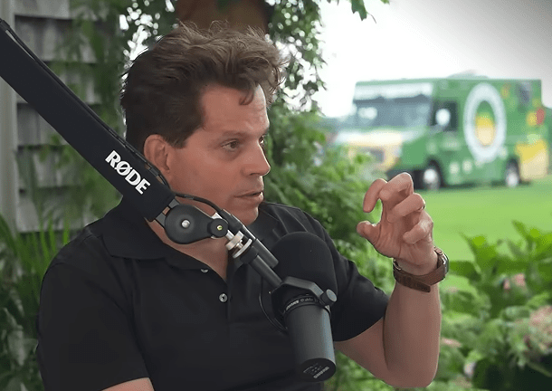 Primer plano de Anthony Scaramucci de perfil, vistiendo una camisa polo negra, hablando frente a un micrófono. Realiza un gesto con la mano izquierda y de fondo se observa vegetación y un vehículo verde desenfocado.