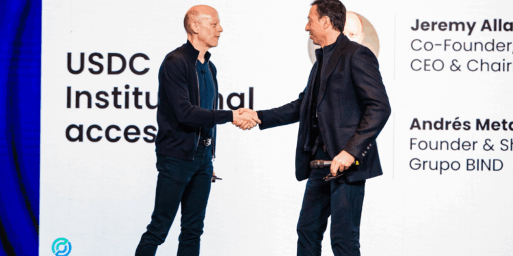 Una fotografía de cuerpo entero de Jeremy Allaire y Andrés Meta estrechándose la mano en un escenario. Ambos hombres llevan chaquetas oscuras y pantalones. Allaire, a la izquierda, es calvo y tiene una mano en su bolsillo trasero. Meta, a la derecha, sonríe y sostiene un micrófono con la mano izquierda.