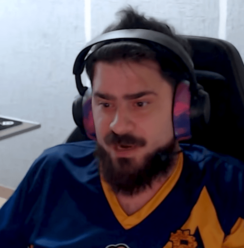 Primer plano de un hombre con barba oscura y pelo despeinado, Daniel Arráez, mirando hacia abajo y a la izquierda con expresión de asombro y concentración. Lleva auriculares negros con copas de color rosa y azul difuminados y una camiseta de fútbol azul oscuro con detalles amarillos. Está sentado en una silla de oficina negra.