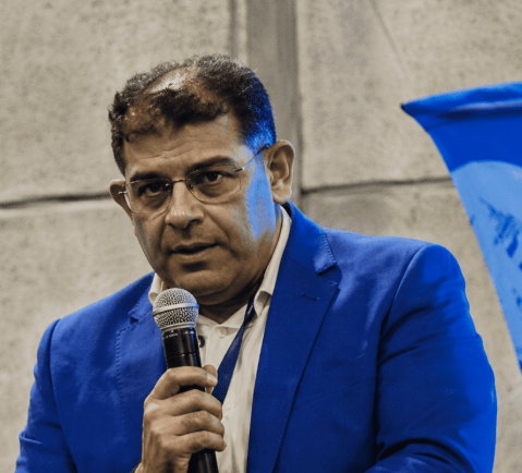 Fotografía de plano medio de Ernesto Portillo, un hombre de mediana edad con gafas y cabello oscuro, vistiendo una chaqueta de traje azul brillante sobre una camisa blanca. Sostiene un micrófono con su mano derecha mientras habla en un evento.