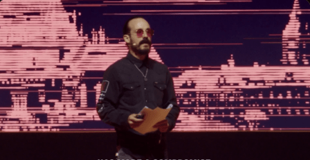 Se observa de medio cuerpo a Francis Pouliot de cabello oscuro recogido, barba y bigote, vistiendo una camisa negra de botones con detalles de costuras y un collar plateado. Lleva puestas unas gafas de sol con lentes tintadas de color rojo y sostiene unos documentos en sus manos en actitud de dar un discurso. Al fondo, se proyecta una pantalla gigante con gráficos digitales pixelados en tonos rojizos que simulan estructuras urbanas abstractas bajo una iluminación baja de escenario.