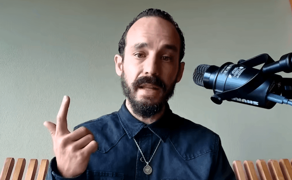 Primer plano de Francis Pouliot, un hombre con cabello oscuro, barba y bigote recortados, vistiendo una camisa oscura y un collar con medalla. Gesticula con su mano derecha levantando el dedo índice mientras habla frente a un micrófono profesional Shure montado en un soporte, con un fondo verde claro.