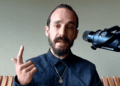 Primer plano de Francis Pouliot, un hombre con cabello oscuro, barba y bigote recortados, vistiendo una camisa oscura y un collar con medalla. Gesticula con su mano derecha levantando el dedo índice mientras habla frente a un micrófono profesional Shure montado en un soporte, con un fondo verde claro.