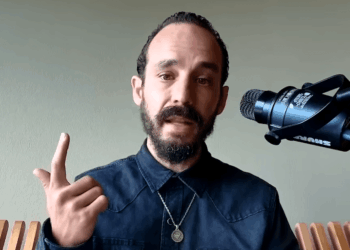 Primer plano de Francis Pouliot, un hombre con cabello oscuro, barba y bigote recortados, vistiendo una camisa oscura y un collar con medalla. Gesticula con su mano derecha levantando el dedo índice mientras habla frente a un micrófono profesional Shure montado en un soporte, con un fondo verde claro.