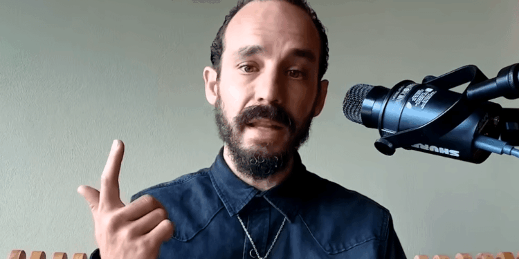 Primer plano de Francis Pouliot, un hombre con cabello oscuro, barba y bigote recortados, vistiendo una camisa oscura y un collar con medalla. Gesticula con su mano derecha levantando el dedo índice mientras habla frente a un micrófono profesional Shure montado en un soporte, con un fondo verde claro.