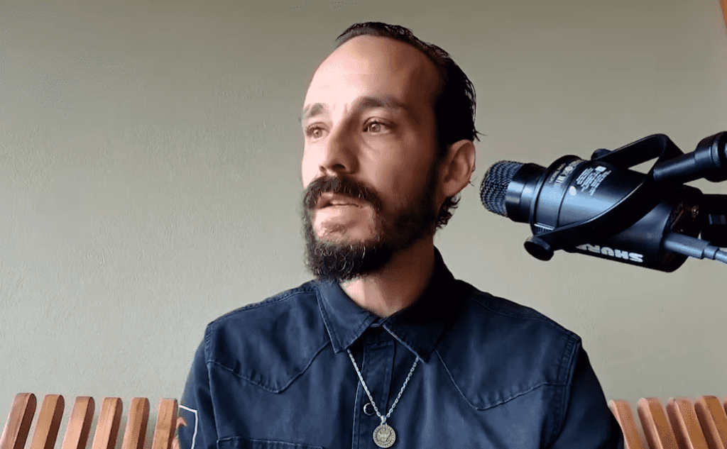 Plano medio en ángulo de tres cuartos de Francis Pouliot mirando hacia la izquierda con expresión seria mientras habla. Se aprecian su cabello oscuro, barba y bigote recortados, una camisa oscura con cuello y una cadena de plata con un dije circular. A la derecha de la imagen se encuentra un micrófono profesional Shure montado sobre un soporte negro, con una pared de color verde claro al fondo.