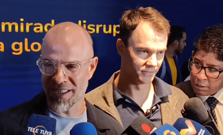 Fotografía de la pantalla mostrando a Román Maniglia y Fred Ehrsam en una entrevista de prensa con múltiples micrófonos, incluyendo Tele Tuya y CNBC. Maniglia sonríe a la cámara, Ehrsam mira hacia abajo. El fondo es azul.