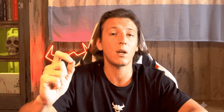 Un plano medio de Micael Margiotta, un hombre de cabello oscuro y lacio hasta los hombros, sentado en una silla de juego blanca y negra. Lleva una camiseta negra con un logo blanco de la cabeza de un toro y hace un gesto con los dedos pulgar e índice unidos para enfatizar un punto mientras mira a la cámara con una expresión seria y la boca ligeramente abierta.