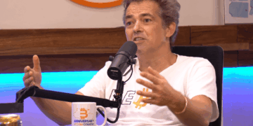 Fotografía de plano medio del activista e informático alemán Michael Ballweg mientras conversa en el podcast "En vivo desde Bitcoin Beach El Salvador". Viste una camiseta blanca abierta sobre una camiseta blanca que lleva impresa la palabra Rebell, (rebelde) escrita con B de Bitcoin.