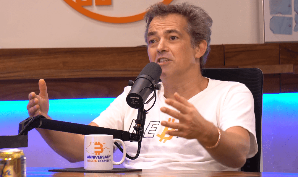 Fotografía de plano medio del activista e informático alemán Michael Ballweg mientras conversa en el podcast "En vivo desde Bitcoin Beach El Salvador". Viste una camiseta blanca abierta sobre una camiseta blanca que lleva impresa la palabra Rebell, (rebelde) escrita con B de Bitcoin.